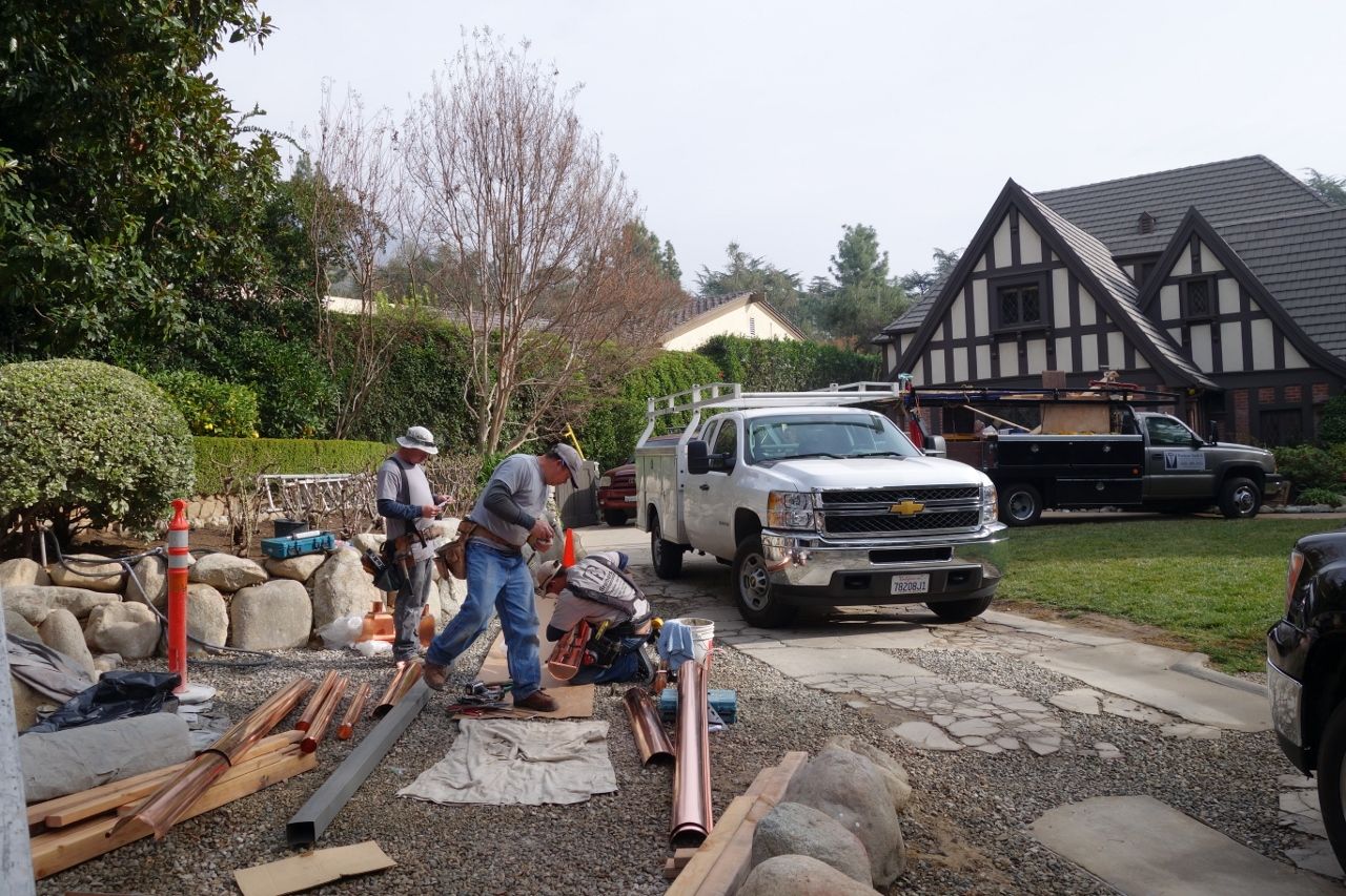 Construction workers near a white truck on a driveway, working on a stone wall. A Tudor-style house is in the background.