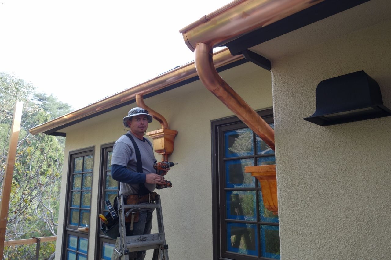 Man on ladder installing copper gutter. House with brown trim.