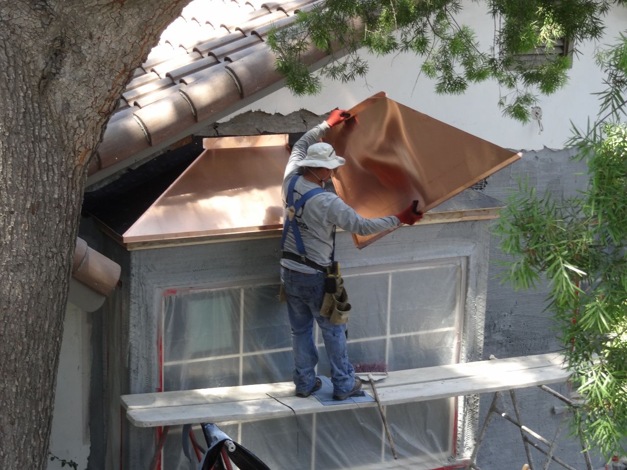 Man installing copper roofing over a window, standing on scaffolding, near a tree.