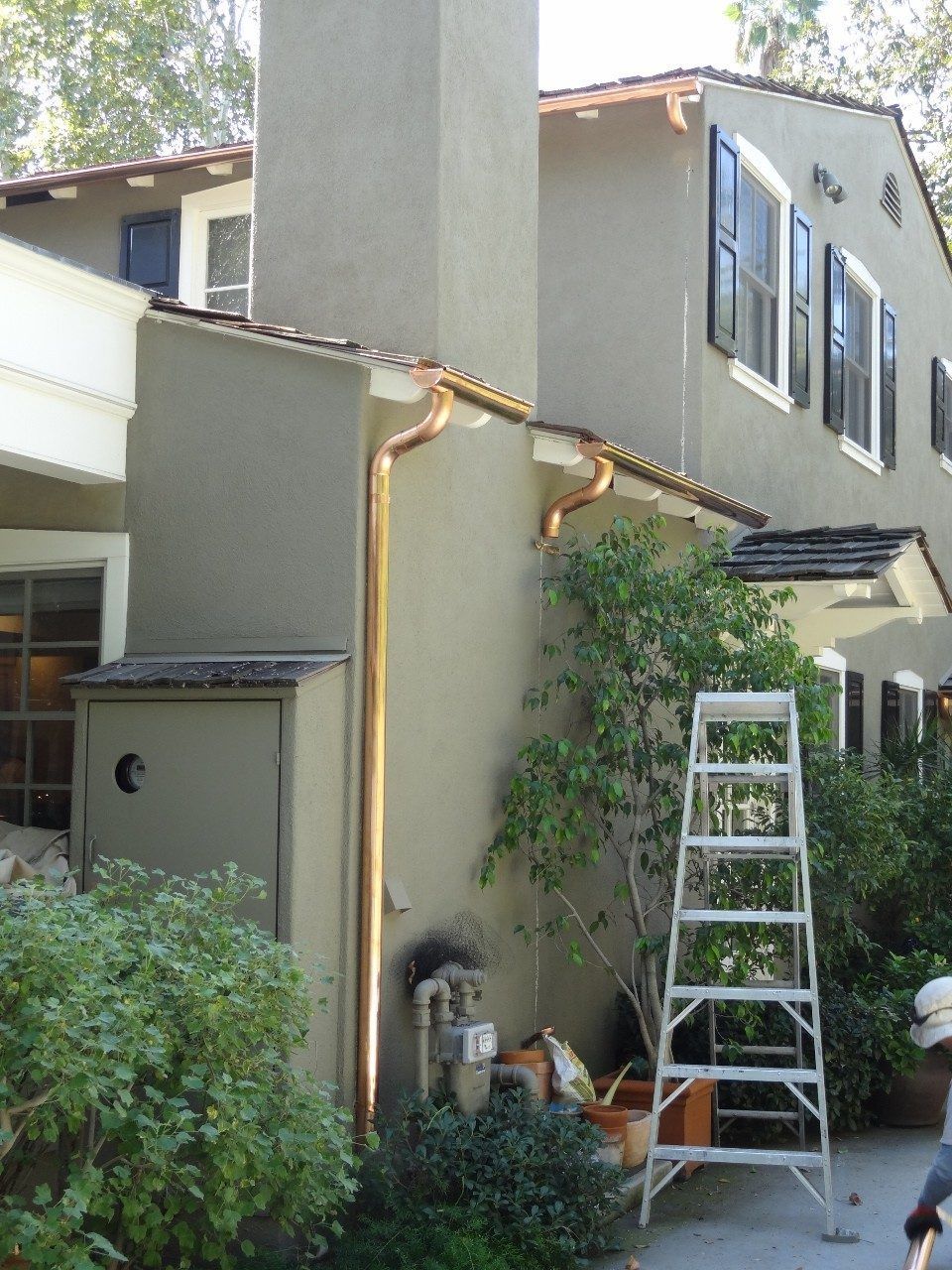 Copper gutters and downspout on a stucco house, with a ladder and greenery in the yard.