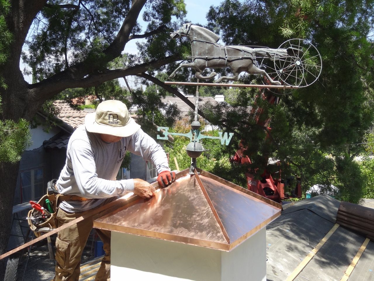Roofer installing copper roof with weathervane. Man wearing hat and tools, on a rooftop in front of a tree.