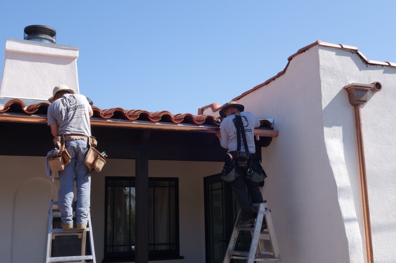 Two roofers installing copper gutters on a white stucco house under a blue sky.