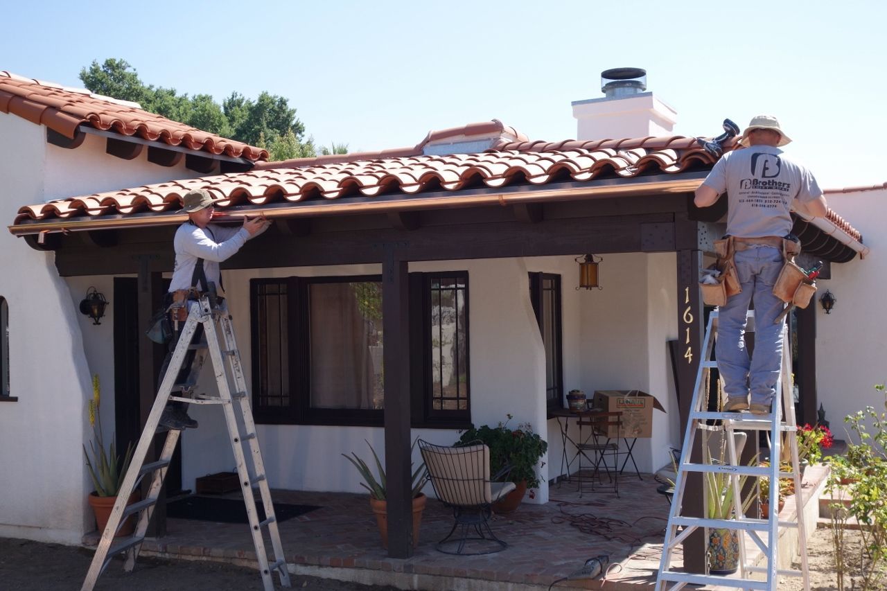 Two workers installing copper gutters on a Spanish-style home with red tile roof. Sunny day.