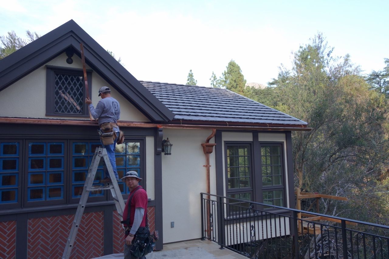 Two workers install copper gutters on a Tudor-style house with dark trim and blue window frames.