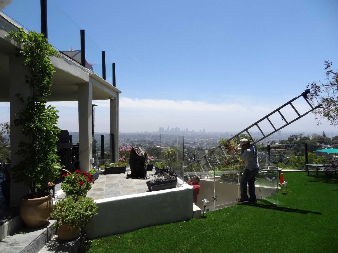 Man carrying ladder on a grassy yard with a city skyline in the background.