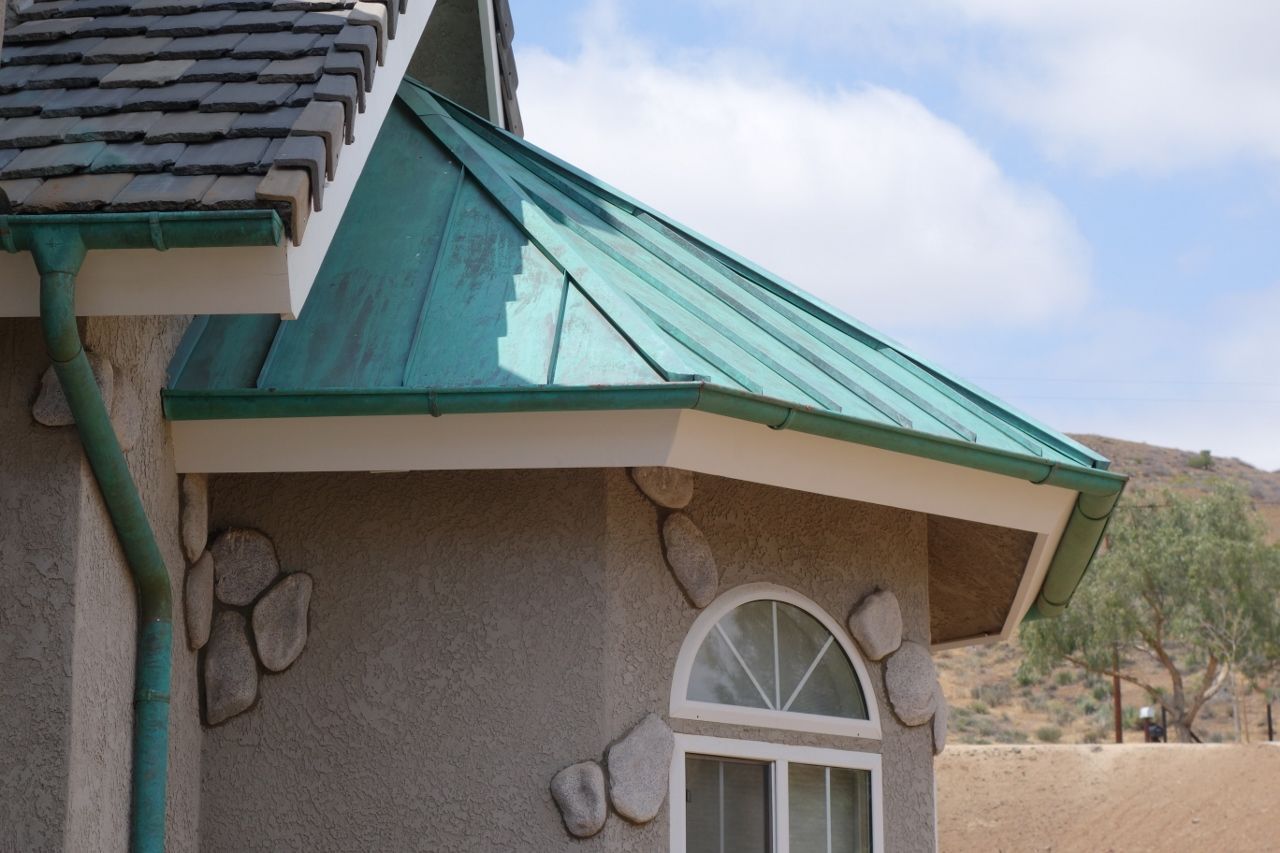 Green metal roof and gutter on a tan house with a stone facade, against a blue sky.