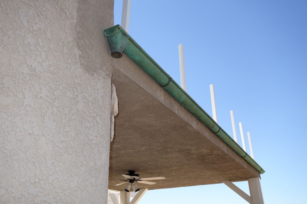 Green gutter on a building's overhang. Exterior with blue sky, stucco wall, and white support posts.