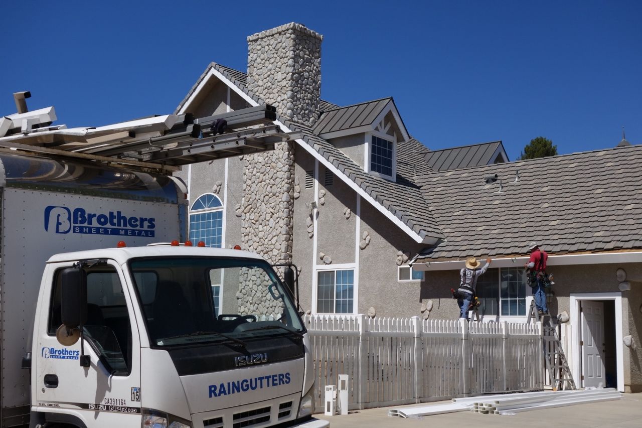 Truck parked in front of a house while workers are installing gutters on the house with blue sky background.