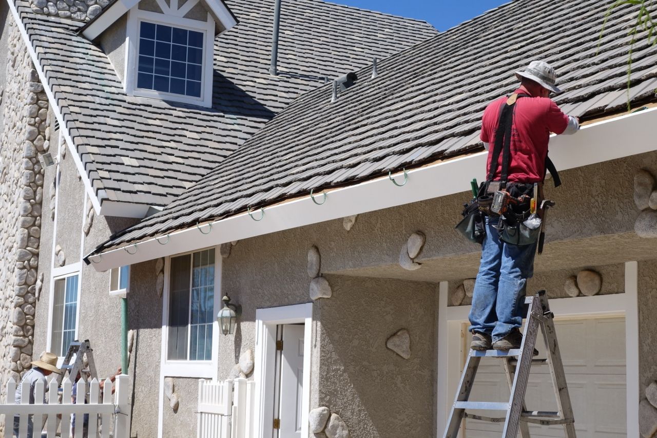 Man on ladder, red shirt, installing gutter on house with stone accents, wood shake roof.
