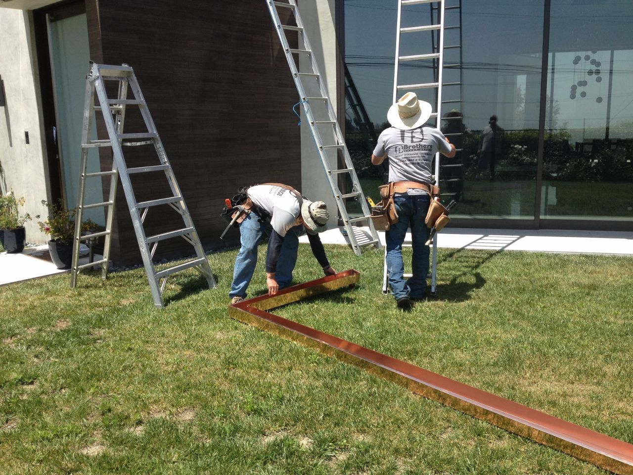 Two workers installing copper gutter on a building's exterior, using ladders, on green grass.