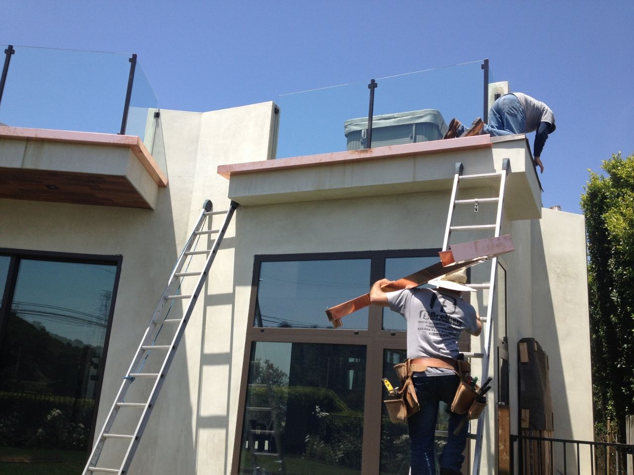 Two workers on a rooftop, carrying materials and working on a building with glass railings on a sunny day.