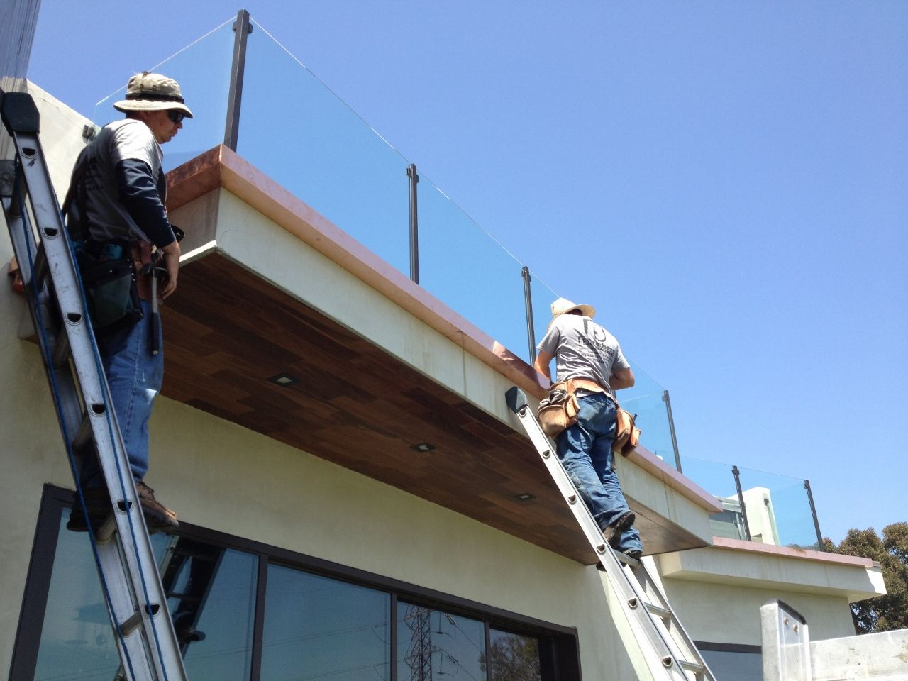 Two workers install glass railing on a rooftop deck, sunny day.