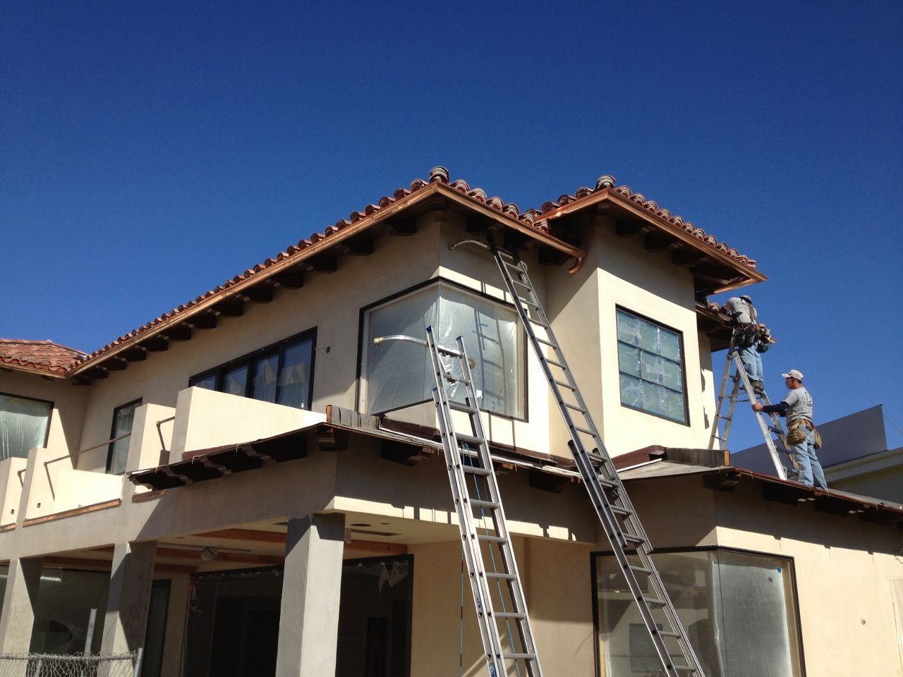 Workers on ladders near the roof of a two-story beige building with a blue sky.