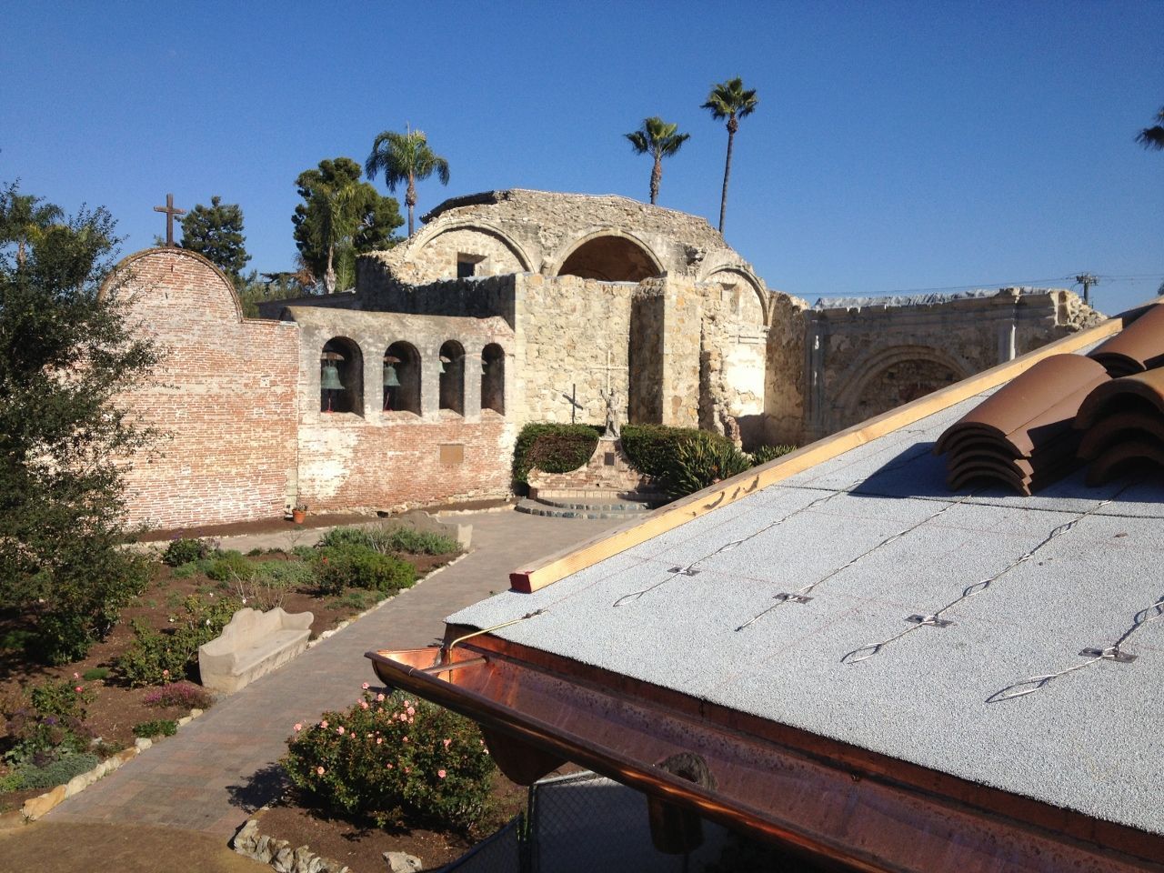 Old mission ruin with brick walls, arched doorways, and a terracotta-tiled roof under a bright blue sky.