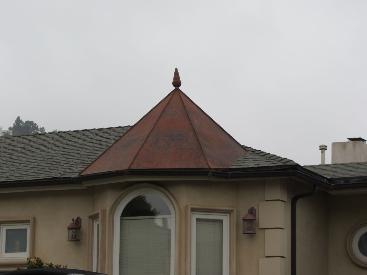 Copper-roofed turret on a house with a decorative finial, gray shingle roof, and beige exterior under a cloudy sky.