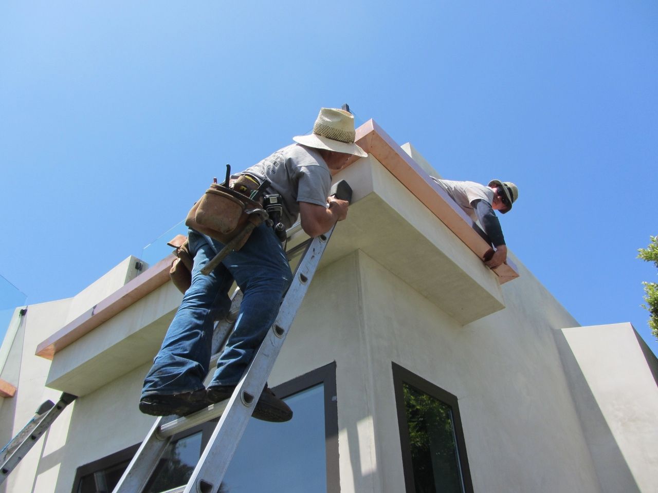 Two construction workers installing a copper-colored gutter on a building's white roof under a clear, blue sky.