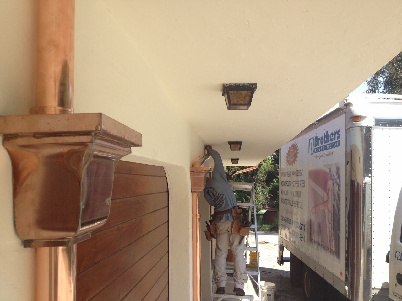 Man on ladder painting stucco under a copper rain gutter, with a truck and garage door.