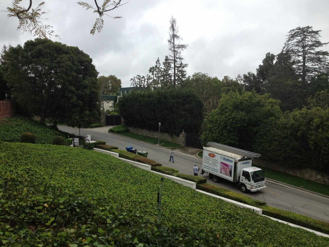Truck on a road surrounded by lush greenery; two figures near the truck. Overcast sky.
