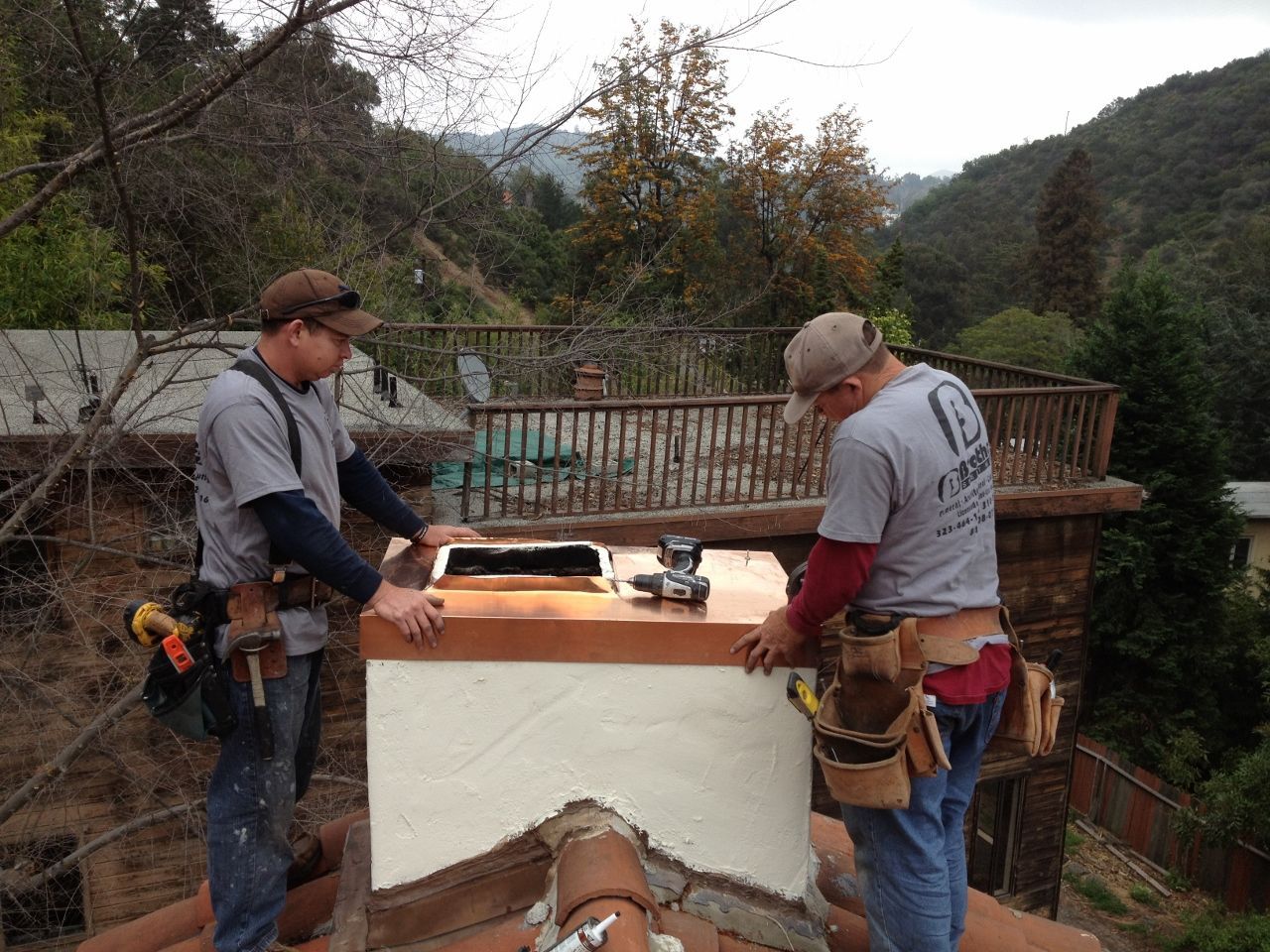 Two roofers installing a copper chimney cap on a brick chimney, with a mountainous backdrop.