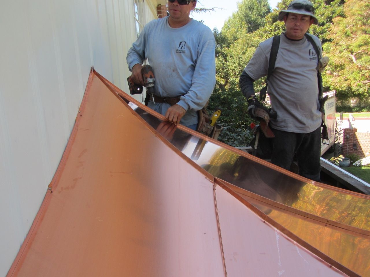 Two men installing copper roofing on a building's edge, using tools.