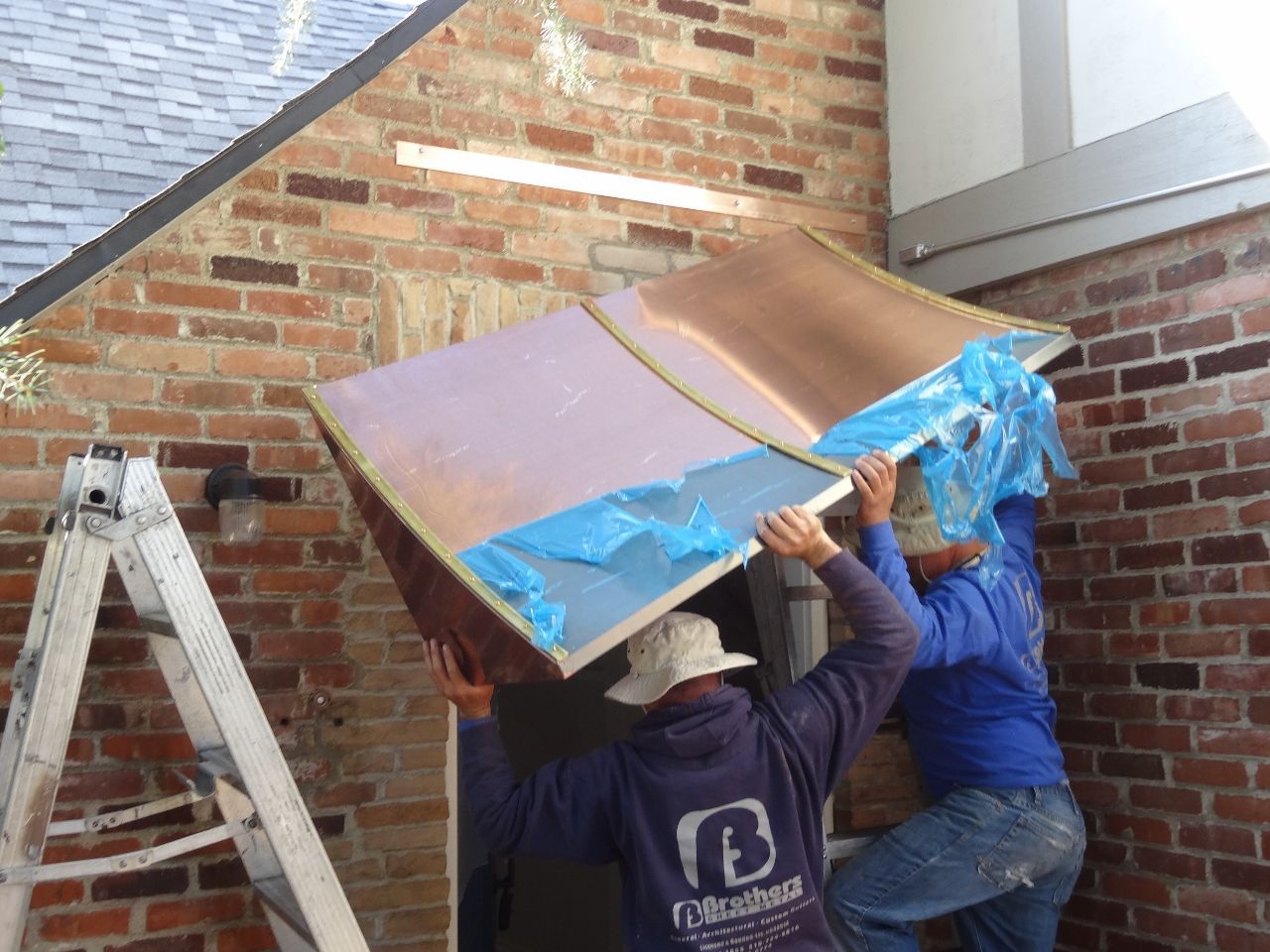 Two workers installing copper panel, brick wall background.