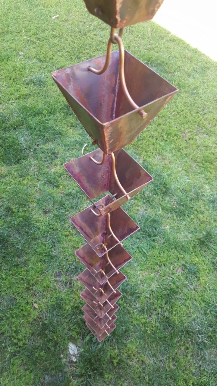 Copper rain chain with square cups hanging against green grass.