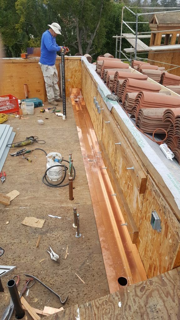 Roofer installing copper flashing on a roof with terracotta tiles; tools and materials scattered around.
