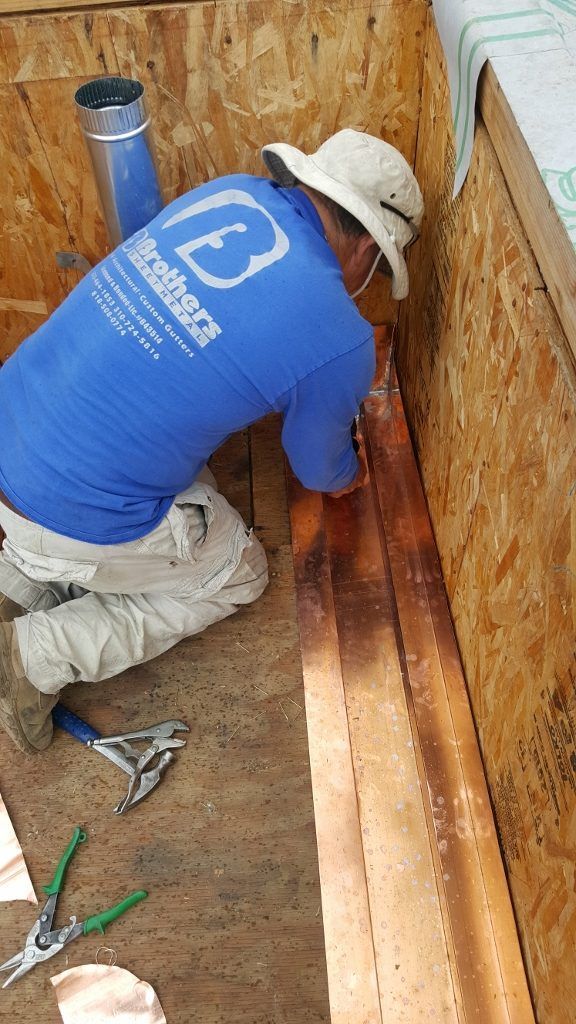 Construction worker installing copper flashing in a roof valley, wearing blue shirt and straw hat.