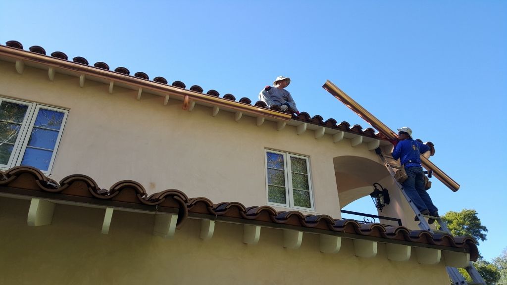Two workers on a roof install a wooden beam. Tan stucco building with red-brown tiles. Blue sky.