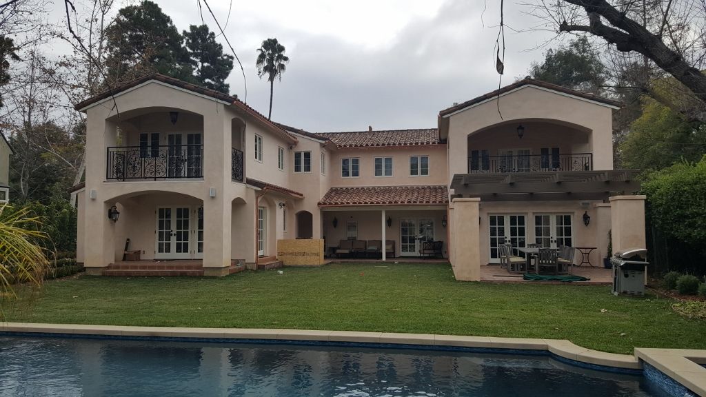 Beige multi-story house with balconies, backyard with green lawn, pool, and cloudy sky.