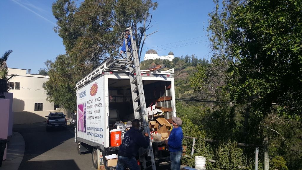 People loading boxes into a moving truck on a sloped street with a tree and a building.