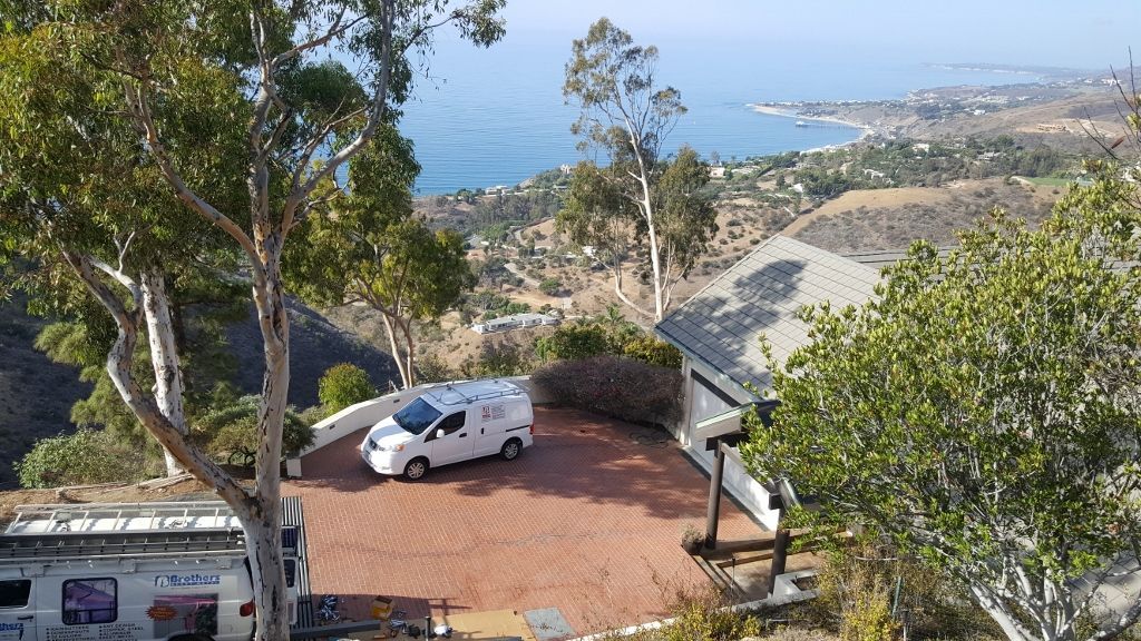 White van parked on a red gravel driveway overlooking a coastal town and ocean, trees in the foreground.