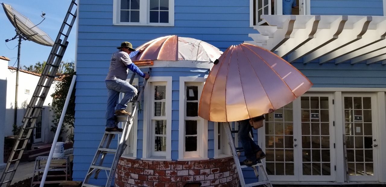 Two workers installing copper dome over windows on a blue house, using ladders.