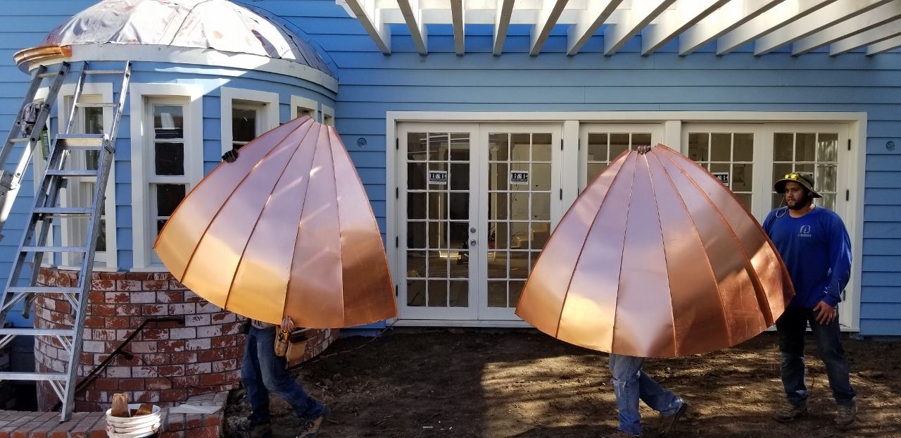 Two workers carrying copper roof pieces near a blue building.