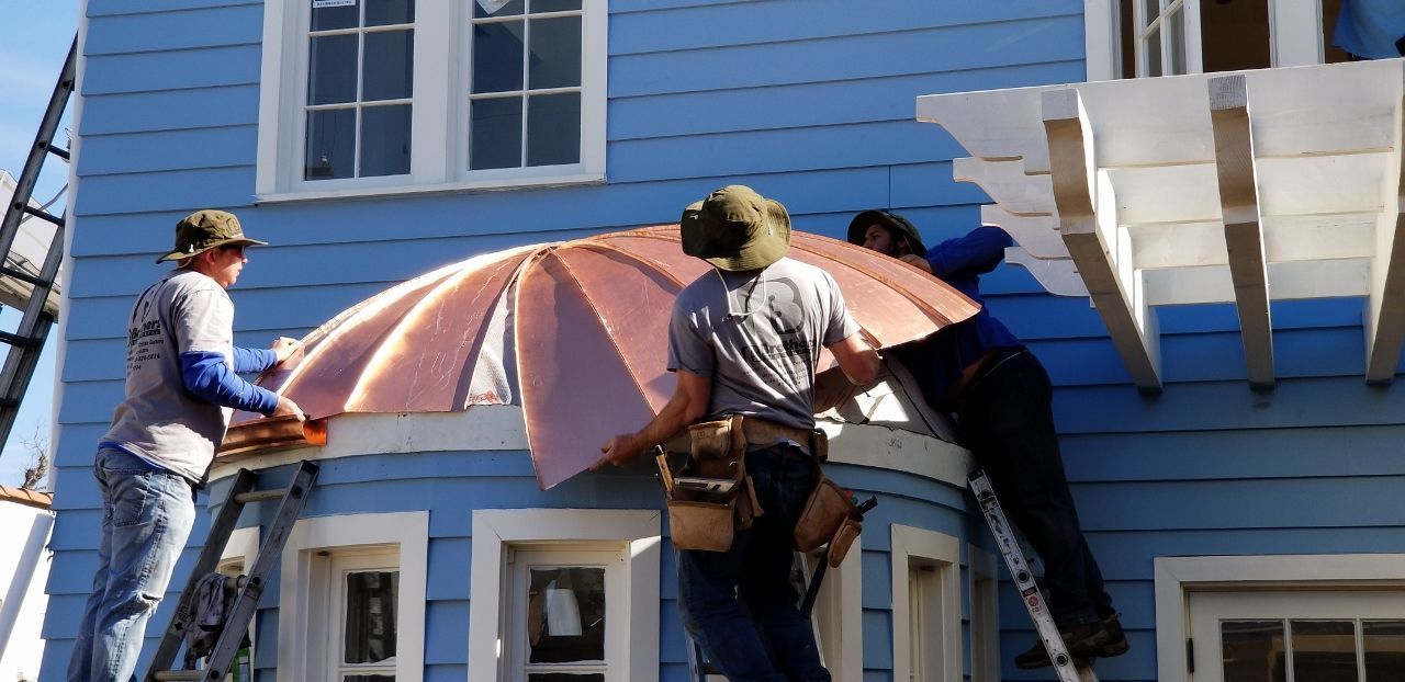 Three workers installing a curved roof awning on a blue house. They are using ladders.