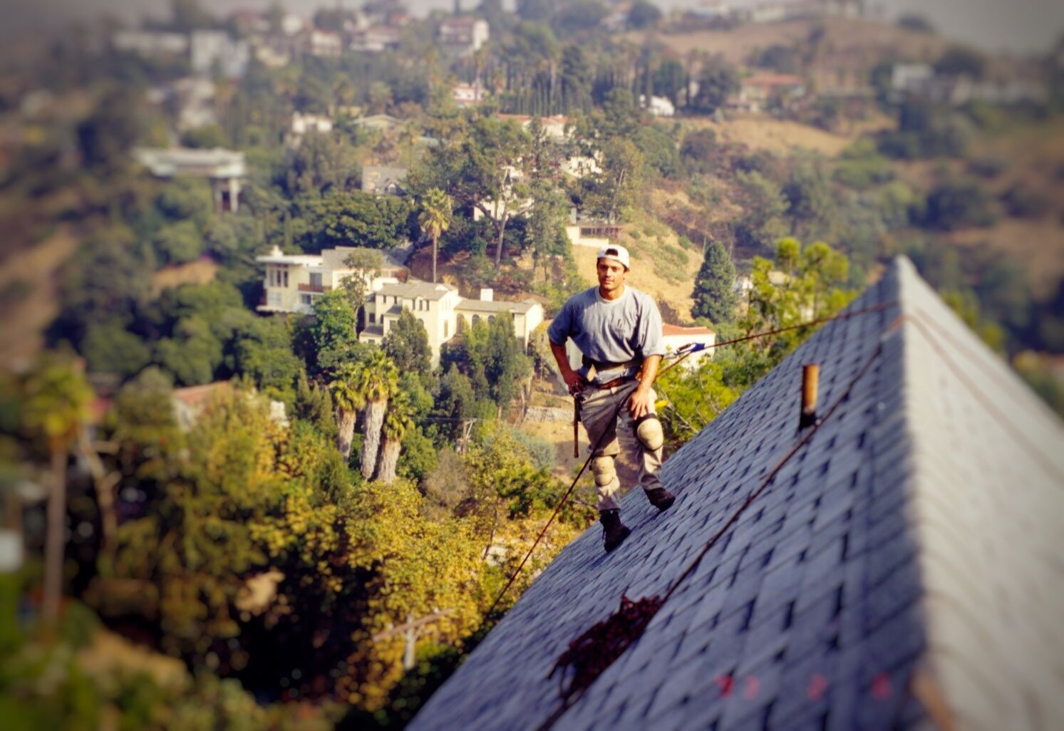 Roofer on a steep roof with a scenic, hillside view. He's wearing work clothes and a hat, surveying the work.