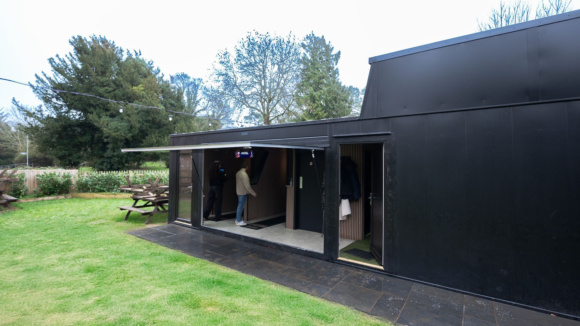 Black converted shipping container with open door, person inside, green lawn, outdoor seating.