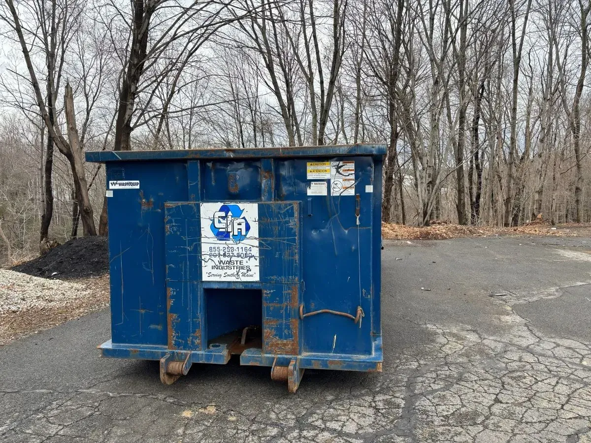 A blue dumpster is sitting on the side of a road in front of trees.