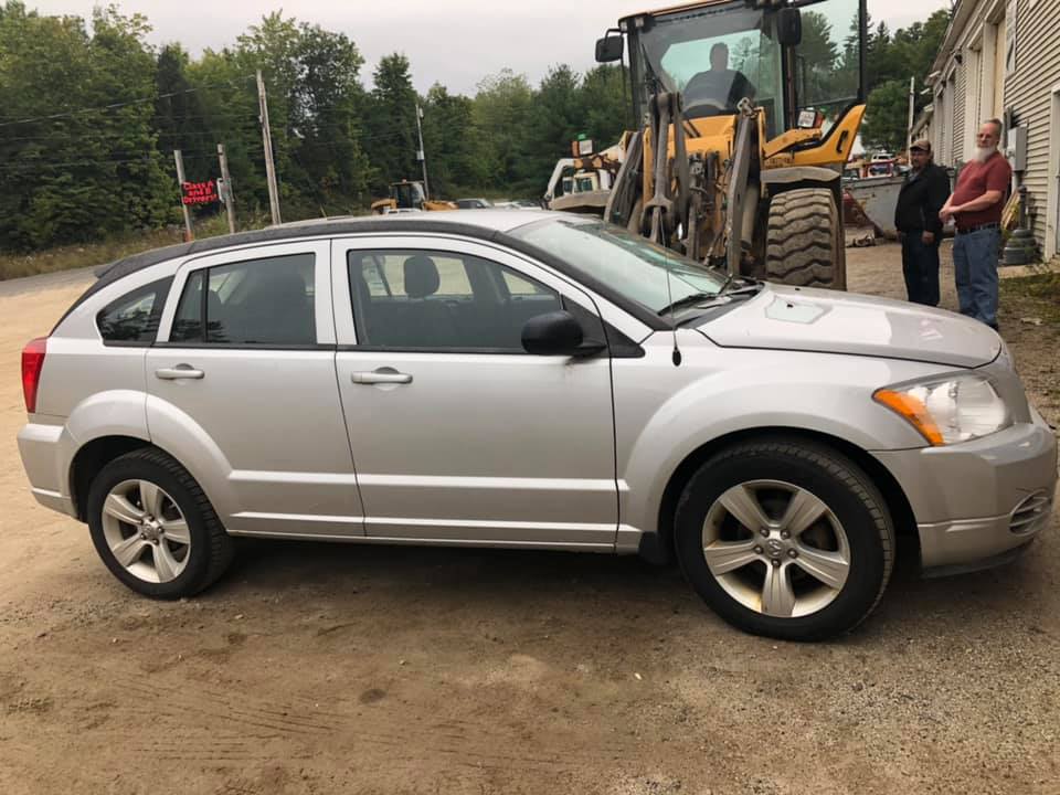 A silver car is parked in a dirt lot next to a bulldozer.