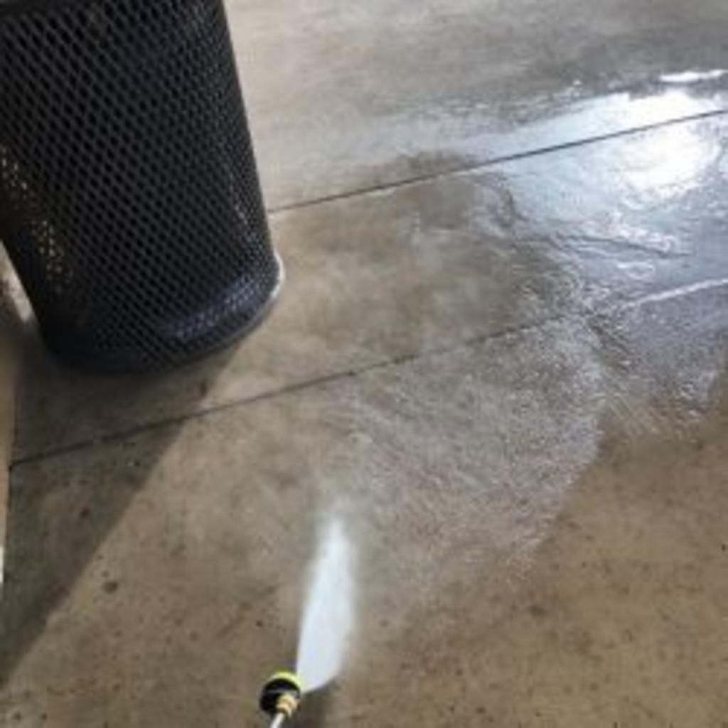A person is using a pressure washer to clean a concrete floor next to a trash can.