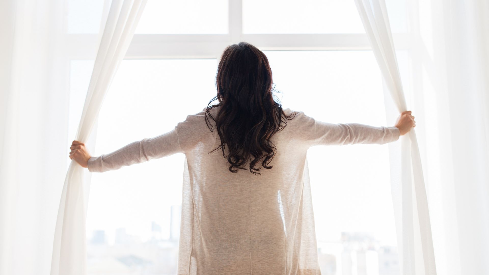 A woman is opening her curtains and looking out of a window.