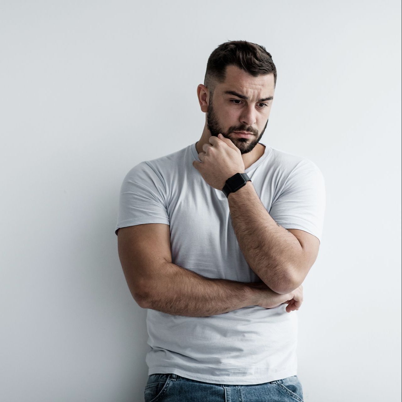 A man with a beard is standing in front of a white wall with his arms crossed and his hand on his chin.