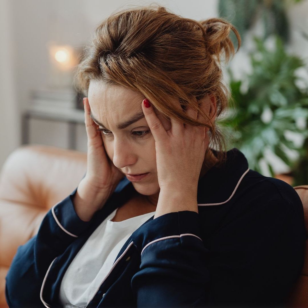 A woman is sitting on a couch with her hands on her head.