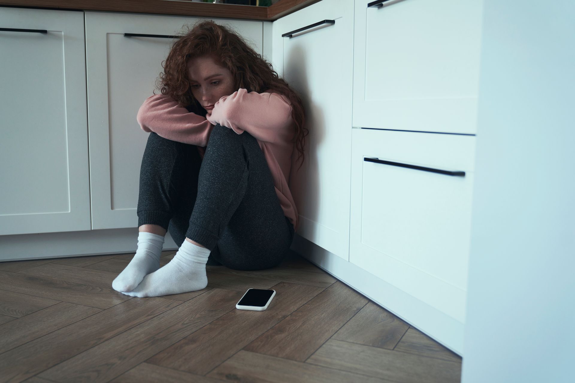 A woman is sitting on the floor in a kitchen next to a cell phone.