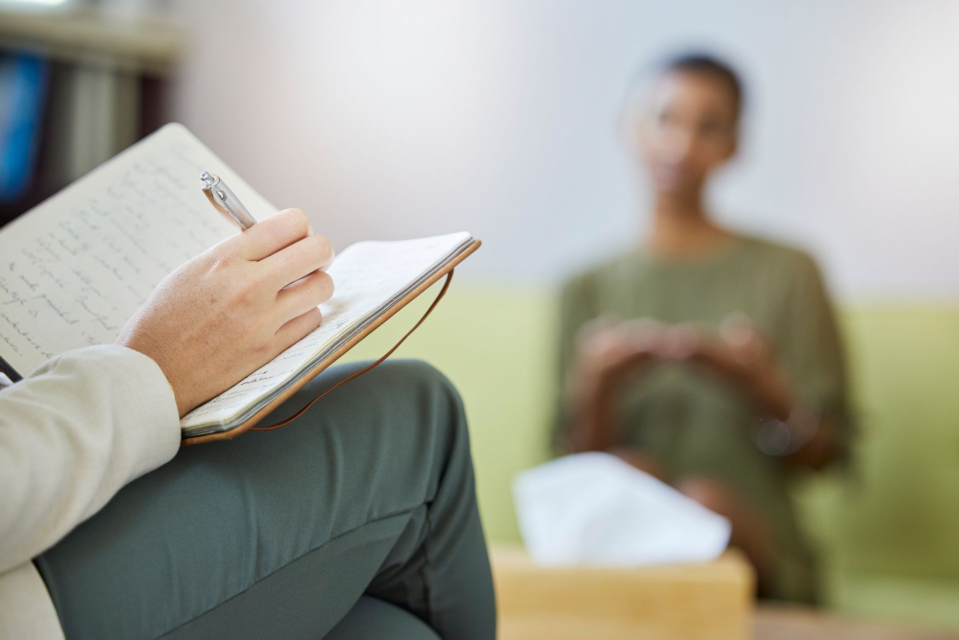 A woman is sitting on a couch talking to another woman while writing in a notebook.