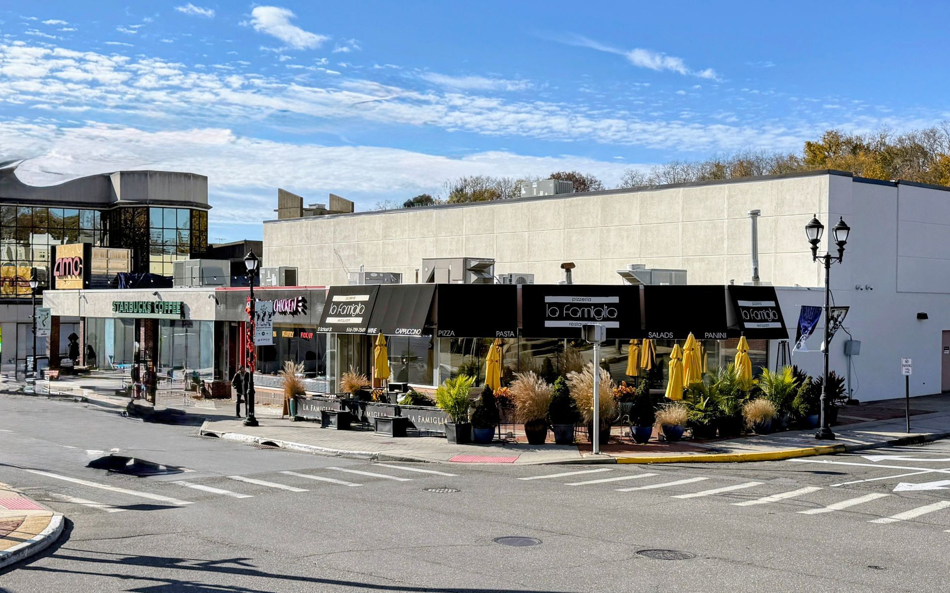 Shops on a corner with awnings, sidewalk seating, and varied signage under a bright, cloudy sky.
