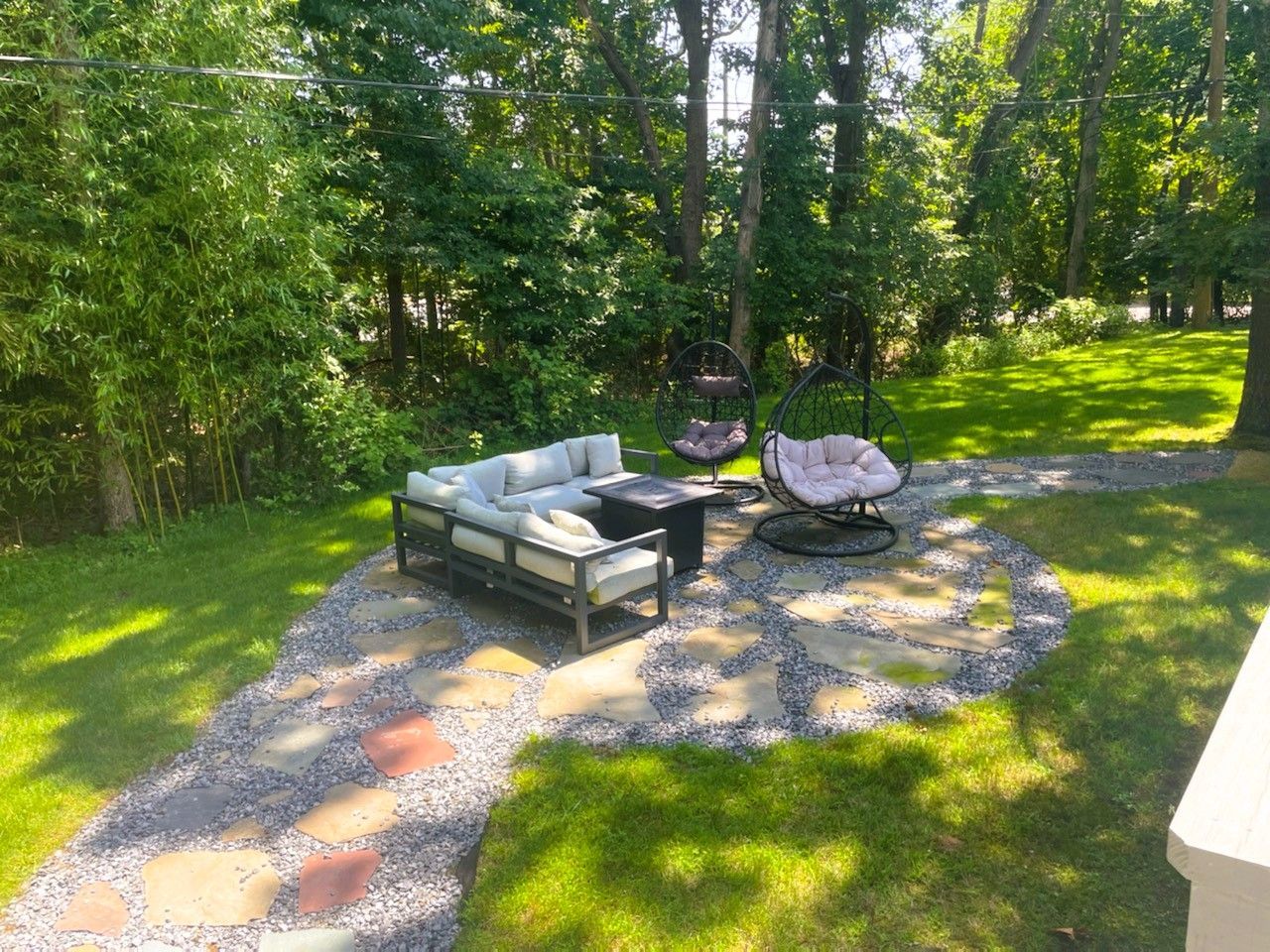 Outdoor patio with gray furniture, gravel path, and hanging chairs in a sunny yard.