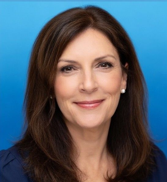 A professional headshot of a person with shoulder-length brown hair, smiling against a solid blue background.