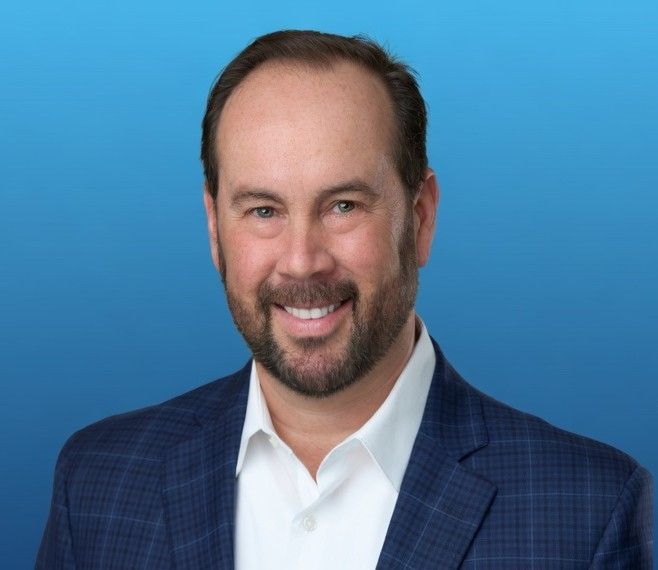 A man with a beard, wearing a patterned blue blazer and white collared shirt, smiles against a plain blue background.