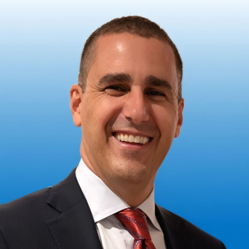 Headshot of a smiling person wearing a dark suit, white shirt, and patterned red tie against a blue gradient background.